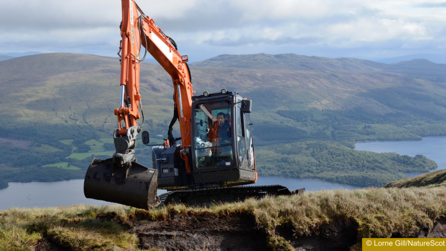 Excavator on a ridge moving peat turf with a lake and mountains in the background