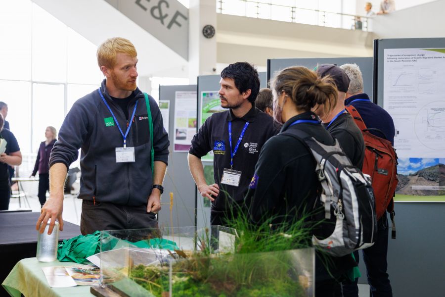 Group of people talking next to a tank of peatland vegetation. There are poster boards in the background
