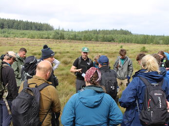 Group of people in outdoor clothing stood listening to a speaker stood in the centre of the group.
