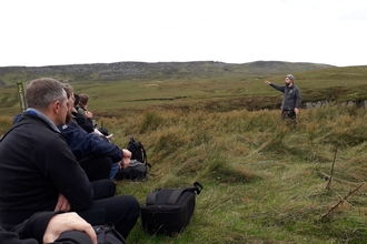 Audience sat on a peatland watching a man pointing and talking
