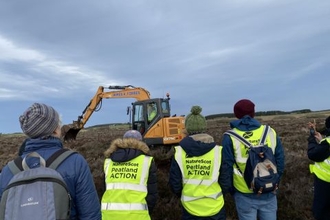Group of people from NatureScot Peatland ACTION watch a machine operator installing peat dams along a peatland ditch.