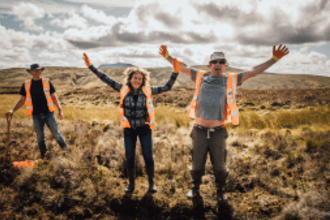 Three SMS volunteers standing on a peatland site. One is holding a spade and the other two are joyfully waving at the camera.