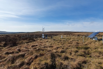 Peatland landscape showing flux tower and solar panel with a blue sky and hills in the background. Image credit Hollie Cooper, UKCEH