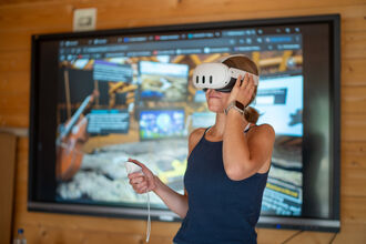 Person wearing a virtual reality headset stood in front of a large screen and holding a hand controller.