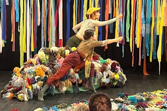 Two performers with their arms outstretched across their bodies. They are stood by a pile of multi-coloured woolen pom-poms with strips of different coloured cloth hanging down behind them.