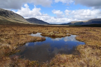 Peatland pool surrounded by mountains. Large clouds in the sky are reflected in the pool.