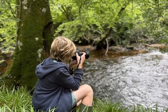 A young boy sitting on a riverbank holding up a camera.