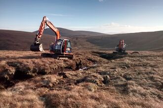 Two diggers on a drained peatland next to a gully. There is blue sky and a mountain in the distance.