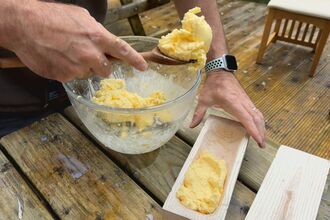 Close-up of someone spooning a thick, creamy yellow mixture from a glass bowl into a wooden dish.