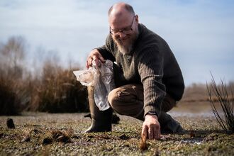 Person in outdoor clothing crouching down holding a bag of plug plants in one hand and pushing a plant into the ground with their other hand.