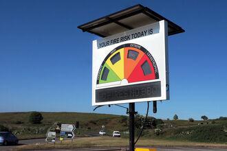 Roadside sign displaying fire risk using a half circle colour coded green, yellow, amber and red. There is a road and vehicles visible in the background.