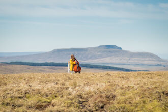 Person in a yellow raincoat sat on a white chair playing a cello on an open moorland. There is a forestry plantation and sloping ridge in the distance.