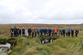 Group of people in outdoor clothing stood behind a bog pool.