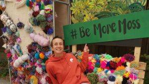 Person smiling and pointing at a sign saying '#BeMoreMoss'. Credit Vicky Akerman