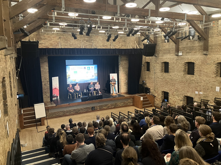 An audience sits in an auditorium listening to a speaker on the stage