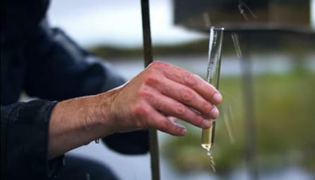 Experiment showing a hand holding a small glass tube collecting a sample of water dripping from a bucket of peat. 
