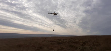 Helicopter flying over a remote peatland restoration site carrying a bucket with materials.