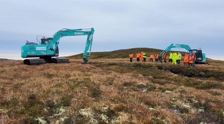 A group of machine operators new to peatland restoration watching an experienced machine operator reprofiling an eroding gully.