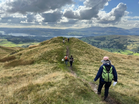 People in outdoor clothing walking away along a moorland path which is high up above a landscape with a large lake and trees in the distance.
