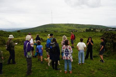 A group of people of mixed ages stood on a grassy area with a green hill in the background.