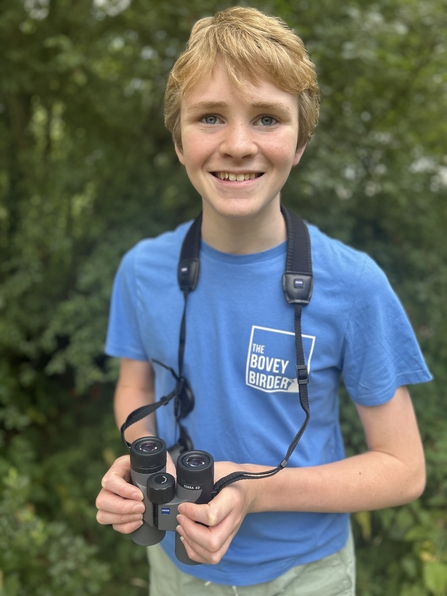 A young boy with a pair of binoculars and a blue t-shirt with 'The Bovey Birder' printed on it.