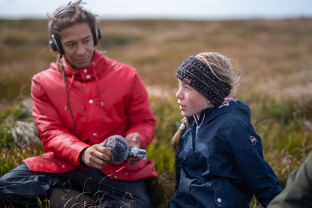 Two people in outdoor clothing sat on a peatland. One is wearing headphones and holding a microphone towards the other.