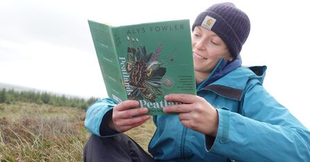 Person in outdoor clothing sat down and holding an open book, displaying a cover with the word 'Peatlands'.