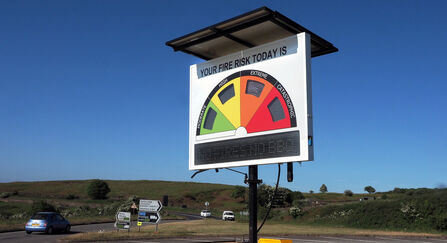 Roadside sign displaying fire risk using a half circle colour coded green, yellow, amber and red. There is a road and vehicles visible in the background.