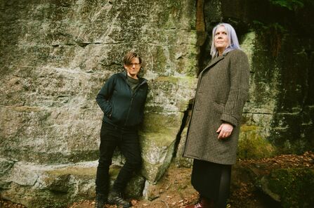 Anna and Clare stood by a stone wall. Clare is leaning against the wall and Anna is near her looking into the distance.
