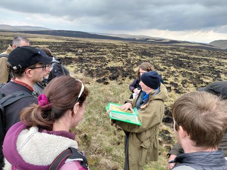 Group of people in outdoor clothing stood around Richard Lindsay, holding aerial images of peatlands.