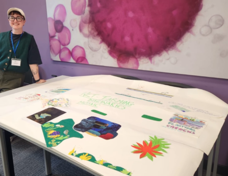 Person stood smiling next to a large sheet of paper covered in individual drawings.
