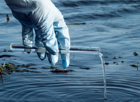 Person's glove-covered hand holding a sample tube over a pool of water
