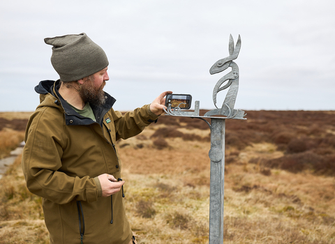 Person in outdoor clothing stood next to a metal sculpture of a hare with a smart phone holder.