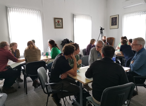 People in casual dress sat in small groups around three tables in a small room