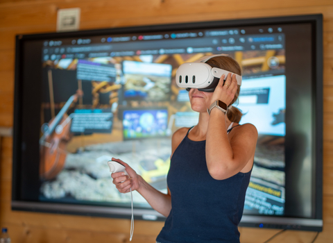 Person wearing a virtual reality headset stood in front of a large screen and holding a hand controller.