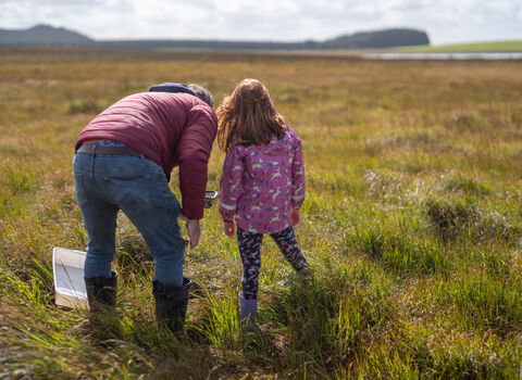 An adult and child stood on a peatland in wellies, looking into a pond net.